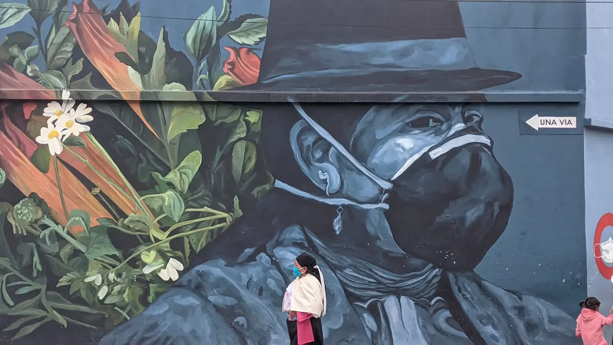 COVID-era street art in Otavalo, Ecuador depicting a masked indigenous woman, while a local resident walks past in the foreground.