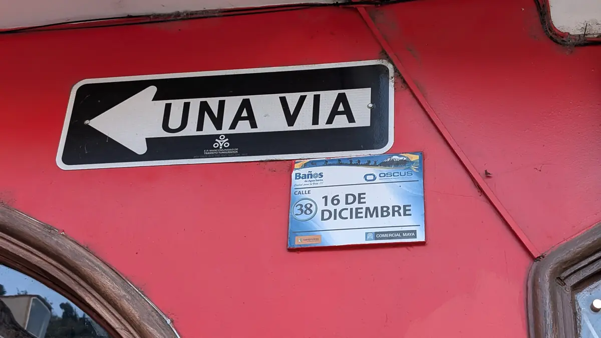 One-way street sign 'Una Via' and street marker for Calle 16 de Diciembre in Baños de Agua Santa, Ecuador.