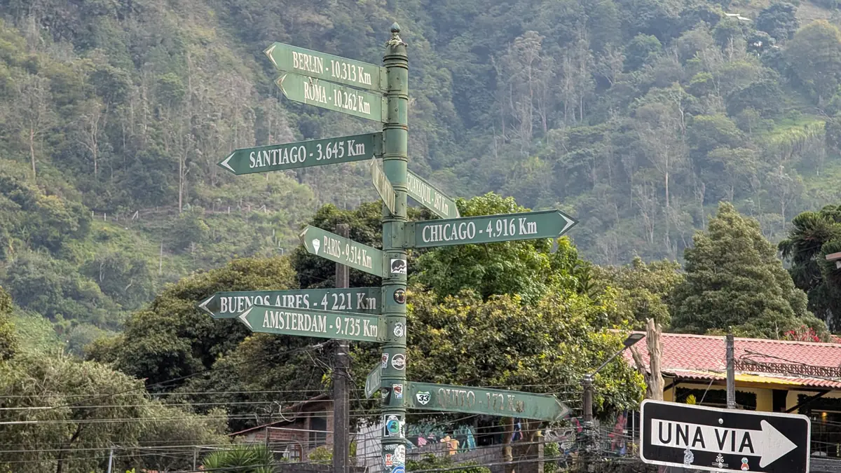 A collection of colorful street and directional signs in the town of Baños de Agua Santa, Ecuador, pointing toward various cities around the world.