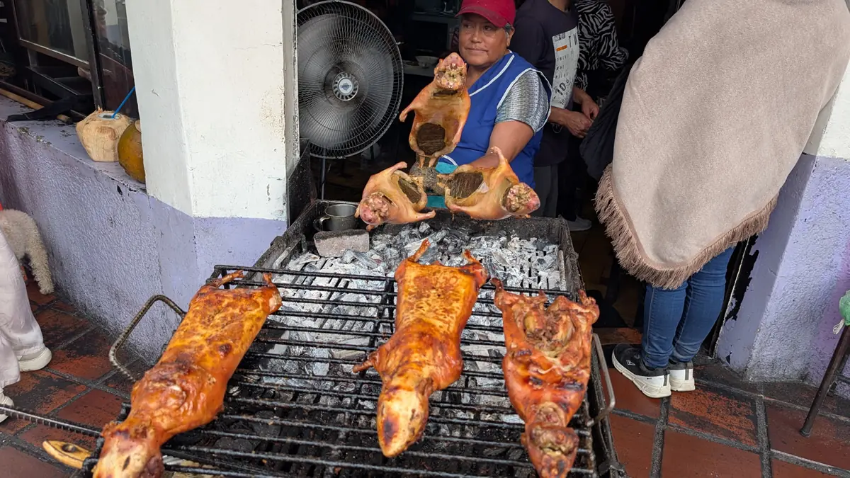 A street food vendor in Baños, Ecuador, preparing roasted cuy (guinea pig) over a charcoal grill on the sidewalk for market customers.